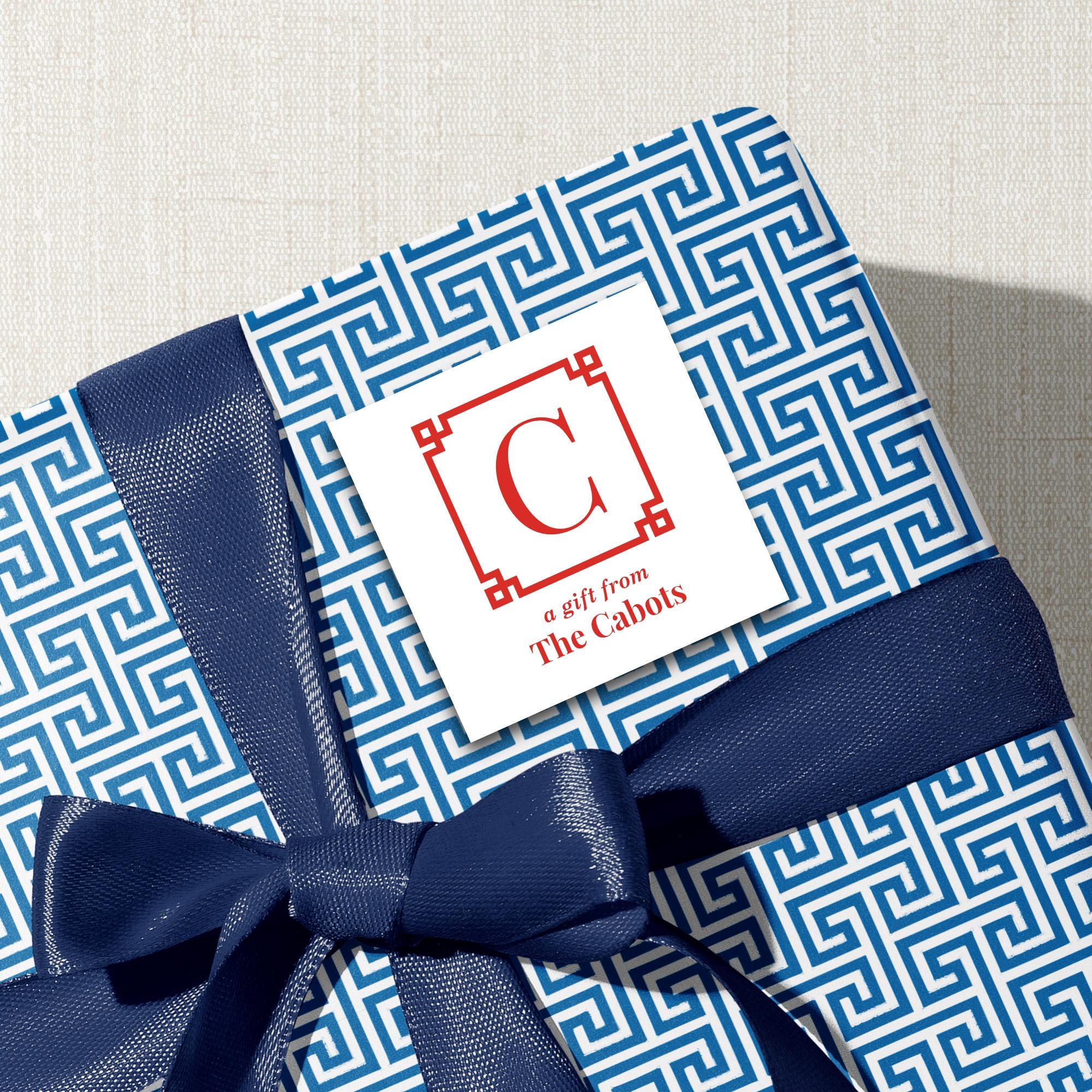A gift wrapped in blue and white patterned paper with a blue ribbon tied around it, featuring a white label with a red letter "C" and text that reads "a gift from The Cabots".