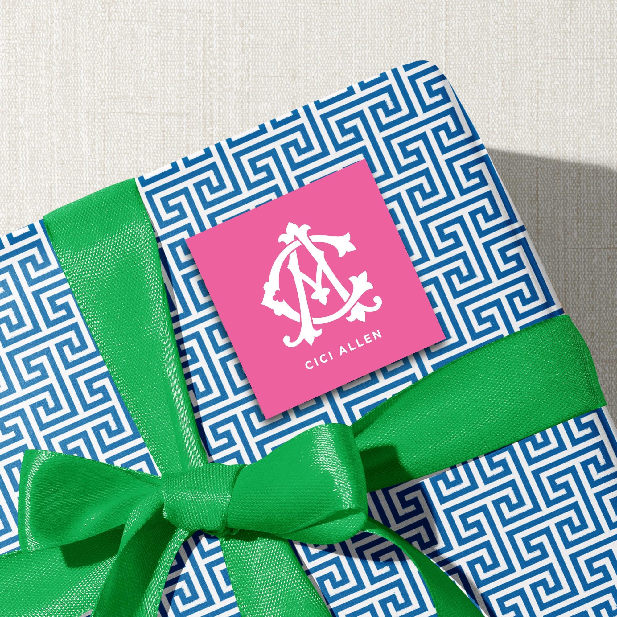 A gift box with a blue and white geometric pattern, tied with a green ribbon, and a pink square sticker with a white monogram logo on it.