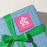 A gift box with a blue and white geometric pattern, tied with a green ribbon, and a pink square sticker with a white monogram logo on it.