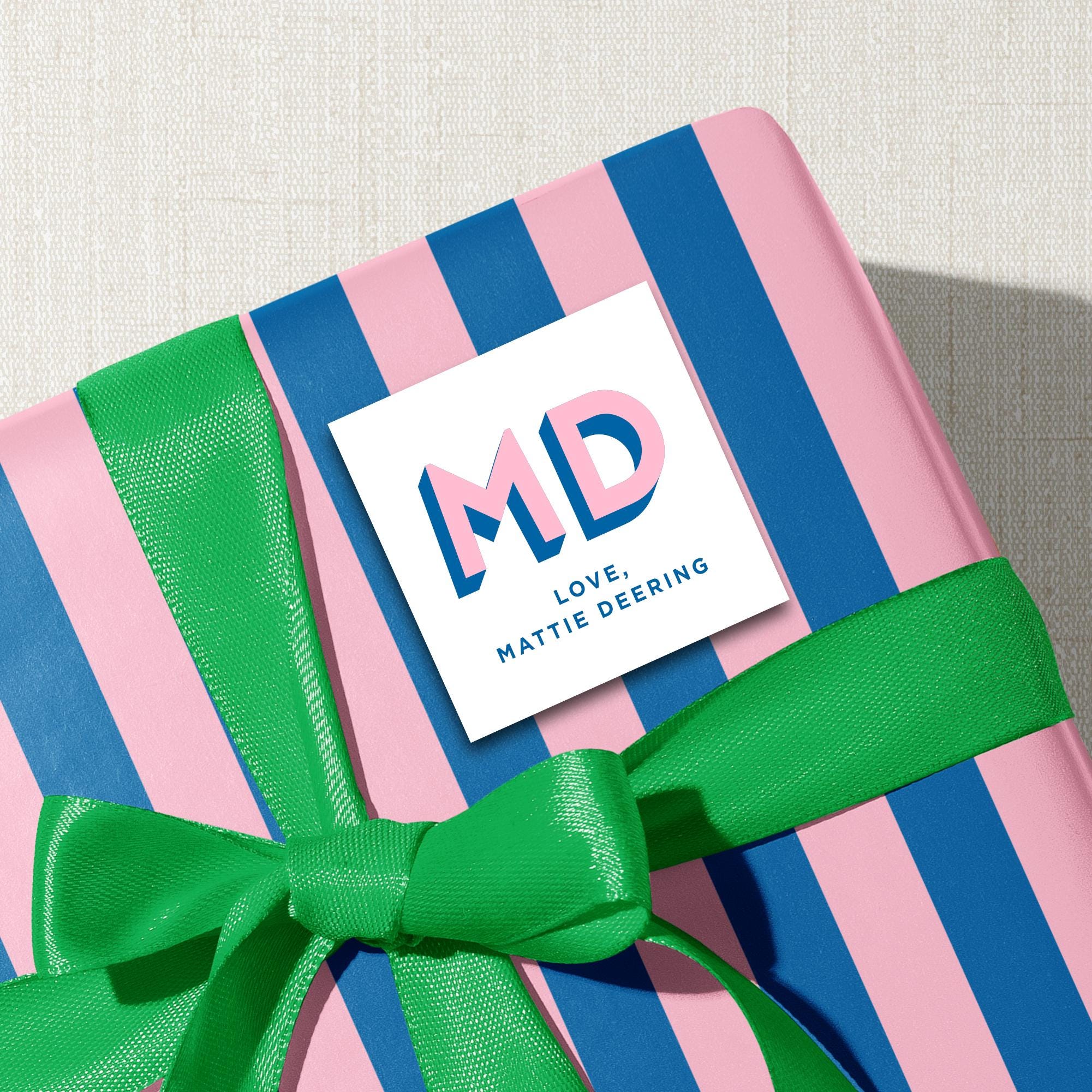 A pink and blue striped gift box with a green ribbon, and a white sticker with the initials 'MD' and the text 'Love Mattie Deering' on it.
