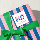 A pink and blue striped gift box with a green ribbon, and a white sticker with the initials 'MD' and the text 'Love Mattie Deering' on it.