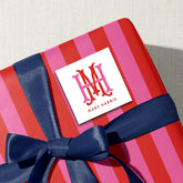A red and pink striped gift box with a blue ribbon, and a white square label with the initials "M.H. M.H." and the name "Mary Harris" printed on it.