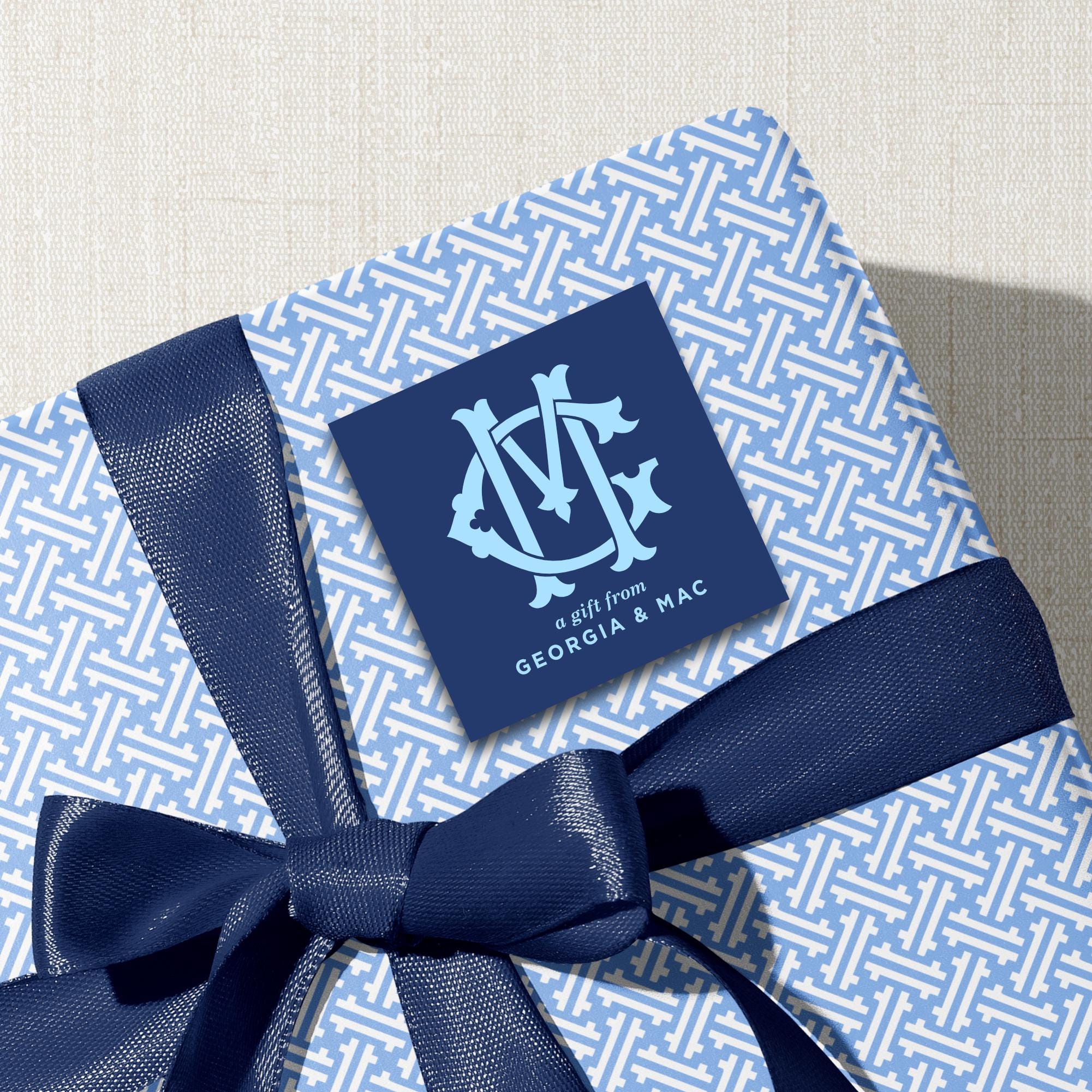 A gift wrapped in blue and white patterned paper with a blue ribbon tied around it. There is a blue square with a monogrammed logo on top of the gift.
