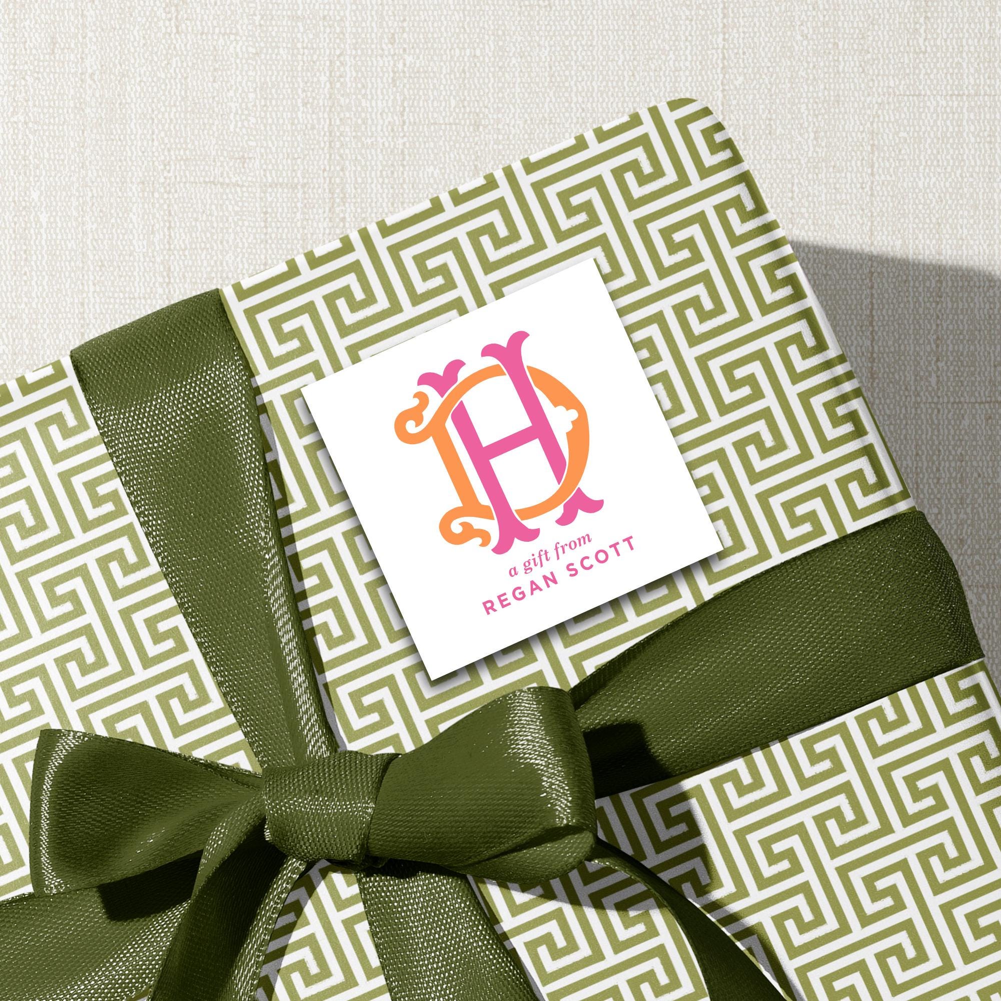 A gift wrapped in a green and white patterned paper with a green ribbon tied around it. There is a white label with a pink and orange logo on the gift.