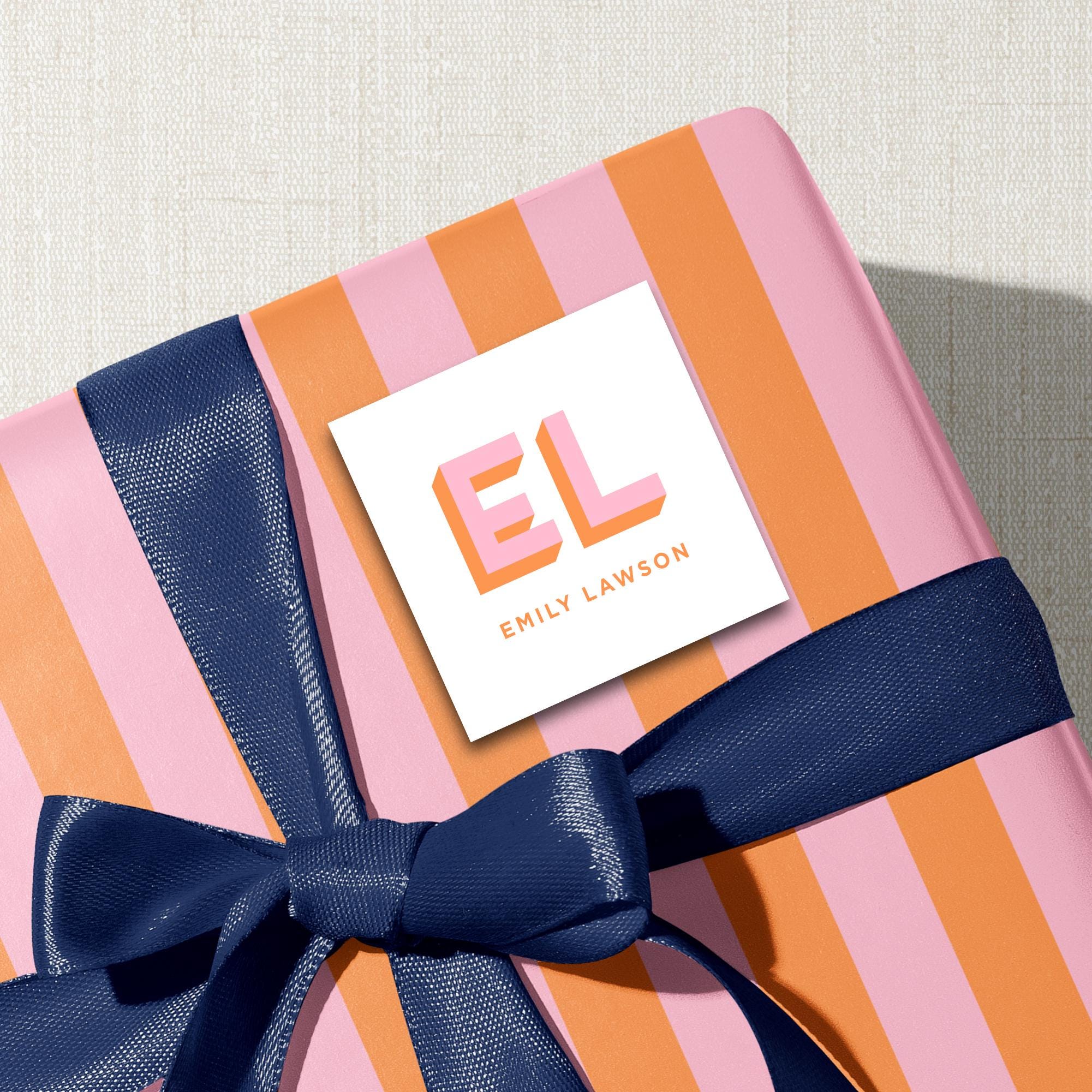 A pink and orange striped gift box with a blue ribbon, and a white label with the text "EL" and "EMILY LAWSON" printed on it.