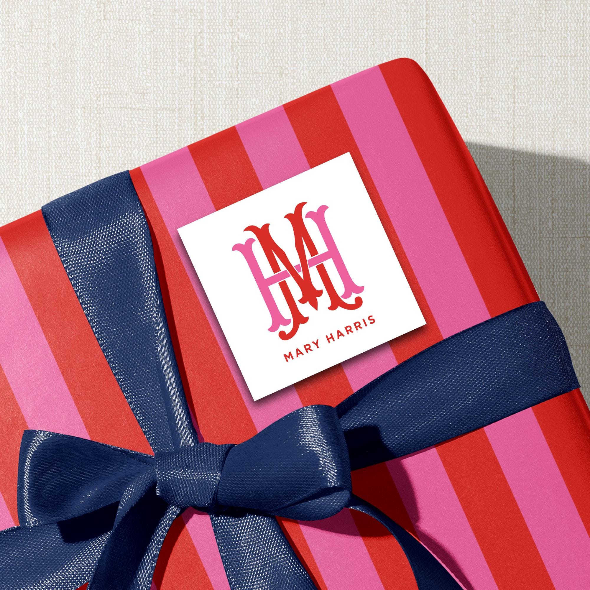 A red and pink striped gift box with a blue ribbon, and a white square label with the initials "M.H. M.H." and the name "Mary Harris" printed on it.